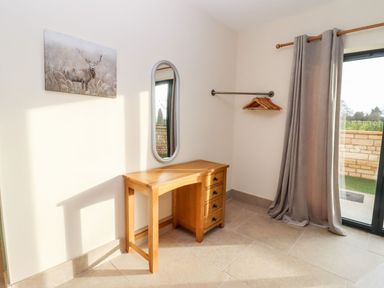 A room corner with a wooden desk, wall mirror, painting of a deer, hanging rail with wooden hangers and a curtain by a glass door at Bears Court Barn 1 Little Rissington near Bourton-On-The-Water