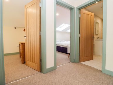 A hallway with three open doors showing a bedroom with skylights a bathroom with a mirror and sink and a room with a chest of drawers at Bears Court Barn 1 Little Rissington near Bourton-On-The-Water