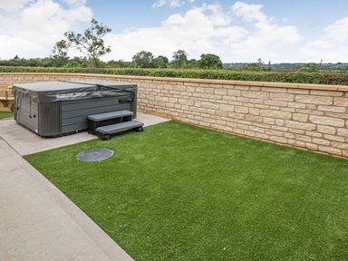 An outdoor area with a covered hot tub steps artificial grass and a stone wall at Bears Court Barn 1 Little Rissington near Bourton-On-The-Water