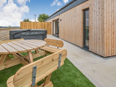 An outdoor patio area with a wooden picnic table and benches a hot tub and wood-paneled building at Bears Court Barn 1 Little Rissington near Bourton-On-The-Water