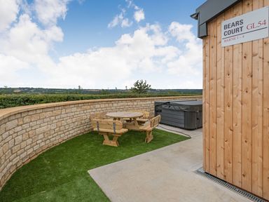 An outdoor seating area with a wooden table and benches next to a stone wall and a hot tub at Bears Court Barn 1 Little Rissington near Bourton-On-The-Water