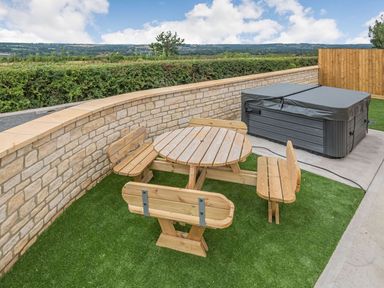 An outdoor seating area with a round wooden table and four benches on artificial grass next to a hot tub at Bears Court Barn 1 in Little Rissington near Bourton-On-The-Water