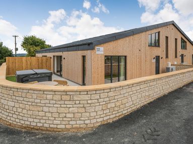 An exterior view of a wooden building with a stone wall and a patio area with table and hot tub at Bears Court Barn 1 Little Rissington near Bourton-On-The-Water