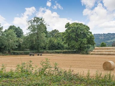 A field with hay bales and a trailer near trees and hills at Bears Court Barn 1 in Little Rissington near Bourton-On-The-Water