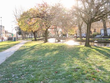 A park with trees and a small stone bridge over a river with ducks and a paved path near buildings at Bears Court Barn 1 Little Rissington near Bourton-On-The-Water