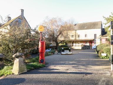 A paved area with a red vintage gas pump near a building and a hedge with a sign saying halt motoring museum ahead at Bears Court Barn 1 Little Rissington near Bourton-On-The-Water