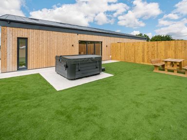 An outdoor area with a hot tub and wooden picnic table surrounded by artificial grass at Bears Court Barn 2 in Little Rissington near Bourton-On-The-Water