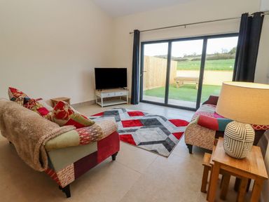 A living room with two patterned sofas a patterned rug a TV on a stand and sliding glass doors leading to a garden at Bears Court Barn 2 Little Rissington near Bourton-On-The-Water