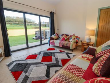 A living room with patchwork sofas a geometric rug and sliding glass doors opening to a garden at Bears Court Barn 2 Little Rissington near Bourton-On-The-Water