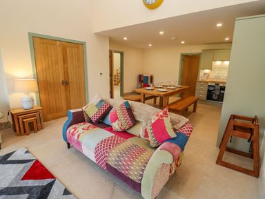 A living room with a patchwork sofa and patterned rug next to a wooden dining table with plates at Bears Court Barn 2 in Little Rissington near Bourton-On-The-Water