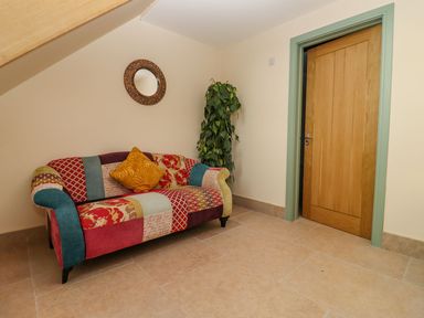 A corner of a room with a multicolored patterned sofa a potted plant a round wall mirror and a wooden door with green trim at Bears Court Barn 2 Little Rissington near Bourton-On-The-Water