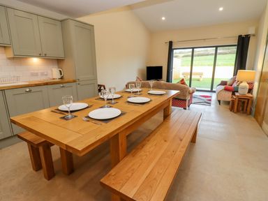A dining area with a wooden table and benches set with plates and glasses next to a kitchen and living room with sofas and a TV at Bears Court Barn 2 Little Rissington near Bourton-On-The-Water