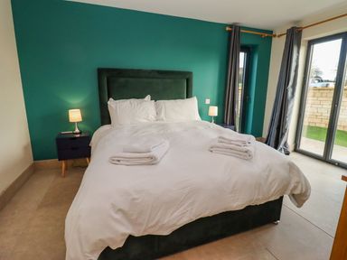 A bedroom with a large bed with white bedding and towels on top two bedside tables with lamps teal accent wall and glass sliding doors with curtains at Bears Court Barn 2 in Little Rissington near Bourton-On-The-Water