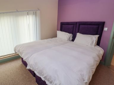 A bedroom with two single beds with white bedding and purple headboards by a window with vertical blinds at Bears Court Barn 2 Little Rissington near Bourton-On-The-Water