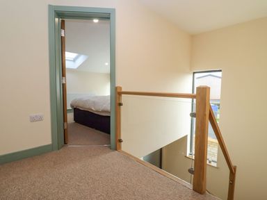 A carpeted hallway with a wooden and glass stair railing leading to a bedroom with a bed near a skylight at Bears Court Barn 2 in Little Rissington near Bourton-On-The-Water