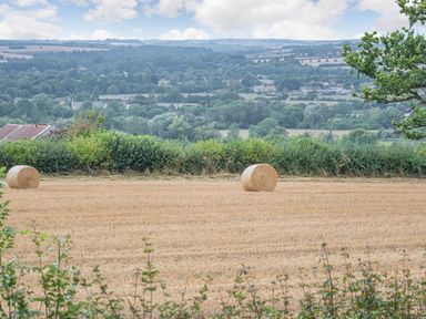 A field with two hay bales and bushes with a rural landscape of trees and houses in the background at Bears Court Barn 2 Little Rissington near Bourton-On-The-Water