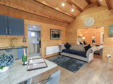 A wooden room with a blue sofa a gray rug blue kitchen cabinets a table with a bottle and flowers and a bed visible in the next room at Drumrane Lodge in Dungiven Londonderry