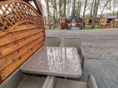 A patio area with four wicker chairs around a glass table and wooden fencing with cabins and trees in the background at Drumrane Lodge in Dungiven Londonderry