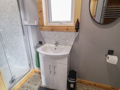 A bathroom with a white sink cabinet under a window a round mirror a shower stall and a toilet paper holder at Railway Lodge in Dungiven Londonderry