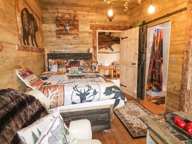 A rustic bedroom with a bed featuring wildlife-themed bedding a wooden table with red apples and a view into a bathroom at Bear Claw Lodge in Castlerock