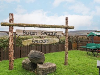 A wooden sign saying Blazin Saddle Saloon on a grassy area with rocks and a fence in the background at Bear Claw Lodge in Castlerock