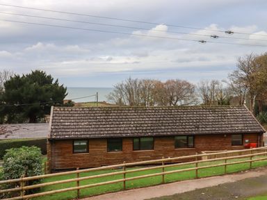 A wooden single-story building with a tiled roof and windows surrounded by grass and trees overlooking the sea at Castle View Lodge in Ilfracombe