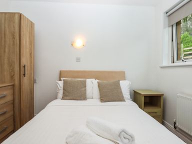 A bedroom with a bed with two brown pillows white linens and folded towels a wooden wardrobe and a bedside table next to a window at Castle View Lodge in Ilfracombe