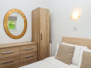 A bedroom with a wooden dresser and wardrobe a bed with cushions a wall light and a round mirror reflecting a window at Castle View Lodge in Ilfracombe