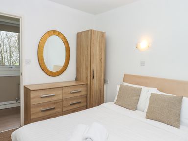 A bedroom with a bed and bedside cushions a wooden wardrobe and dresser with a round mirror on the wall at Castle View Lodge in Ilfracombe