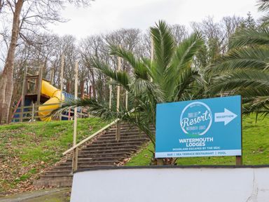 A set of wooden stairs leading up a hill with a playground slide and a blue reception sign for Watermouth Lodges surrounded by trees and palm plants at Castle View Lodge in Ilfracombe