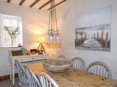 A dining room with a wooden table and chairs a side table with a lamp books and flowers and a coastal painting on the wall at Riverwash Cottage in Dwygyfylchi