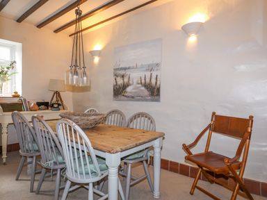 A dining room with a wooden table surrounded by six gray chairs a wooden and leather folding chair a sideboard with books and a lamp and a beach-themed painting on the wall at Riverwash Cottage in Dwygyfylchi