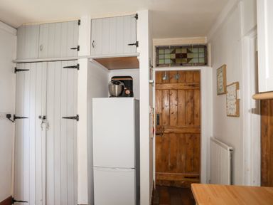 A kitchen area with white cupboards a refrigerator with a mixer on top a wooden door with stained glass above and framed wall art at Riverwash Cottage in Dwygyfylchi