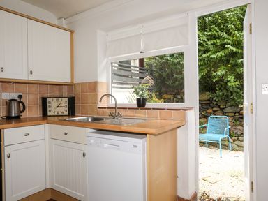 A kitchen with white cabinets a wooden countertop a sink a clock a kettle and a window with a plant outside and an open door to a garden with a blue chair at Riverwash Cottage in Dwygyfylchi