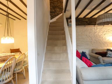 A carpeted staircase between a dining area with wooden chairs and a living room with sofas and cushions at Riverwash Cottage in Dwygyfylchi