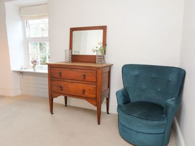 A corner of a room with a wooden chest of drawers with a mirror and vases on top next to a blue upholstered chair at Riverwash Cottage in Dwygyfylchi