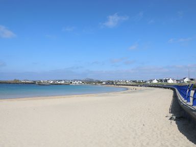 A sandy beach with a blue railing along the pathway and houses in the distance at Seaddler in Trearddur Bay