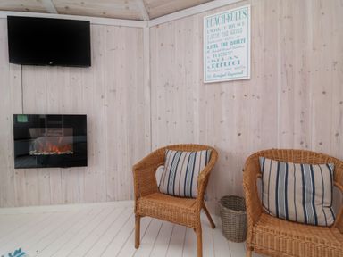 A room with two wicker chairs with striped cushions a wall-mounted electric fireplace a wall-mounted tv and a beach rules sign at Shipload Bay near Hartland