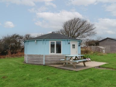 A small octagonal light blue cabin with a picnic table on a wooden deck in a grassy area at Shipload Bay near Hartland