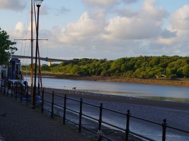A riverside walkway with a boat docked next to a railing and a bridge in the background at Shipload Bay near Hartland