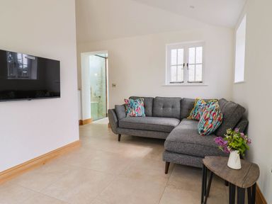 A living room with a grey sectional sofa with colorful pillows a small wooden side table with flowers and a wall-mounted flat-screen TV at Upper Cottage in Rocombe near Uplyme