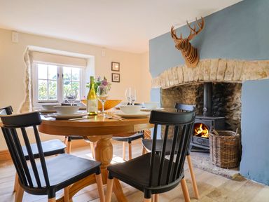 A dining room with a round wooden table set with plates glasses and a bottle near a fireplace with a wood burning stove at Upper Cottage in Rocombe near Uplyme