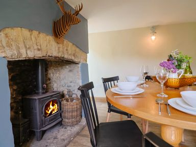 A dining area with a round wooden table set with white bowls plates and wine glasses a wood burning stove with fire in a stone fireplace at Upper Cottage in Rocombe near Uplyme