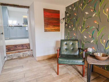 A corner of a room with a green armchair next to a wooden table with a bowl and a wall with floral wallpaper at Upper Cottage in Rocombe near Uplyme