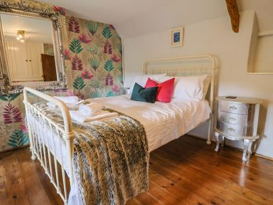 A bedroom with a metal bed frame with white bedding and colorful pillows a fur throw a floral wallpaper and a silver bedside table at Upper Cottage in Rocombe near Uplyme