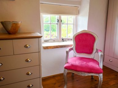 A room with a pink upholstered chair near a window and a chest of drawers with a bowl on top at Upper Cottage in Rocombe near Uplyme