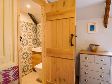 A bathroom with patterned tiles and a wooden vanity seen through a wooden door next to a chest of drawers with a bowl and framed picture at Upper Cottage in Rocombe near Uplyme