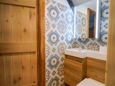 A bathroom with a wooden door a patterned tiled wall a white sink with wooden cabinets and a mirror at Upper Cottage in Rocombe near Uplyme