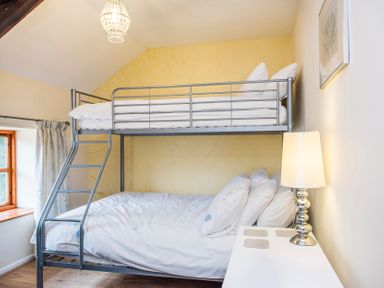 A bedroom with a metal bunk bed a bedside table with a lamp and a window with curtains at Mill Cottage in Aylesbeare