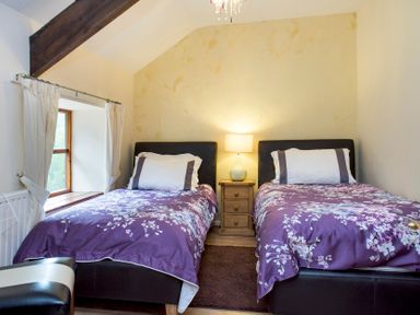 A bedroom with two single beds with floral purple bedspreads a wooden nightstand with a lamp and a window with white curtains at Mill Cottage in Aylesbeare